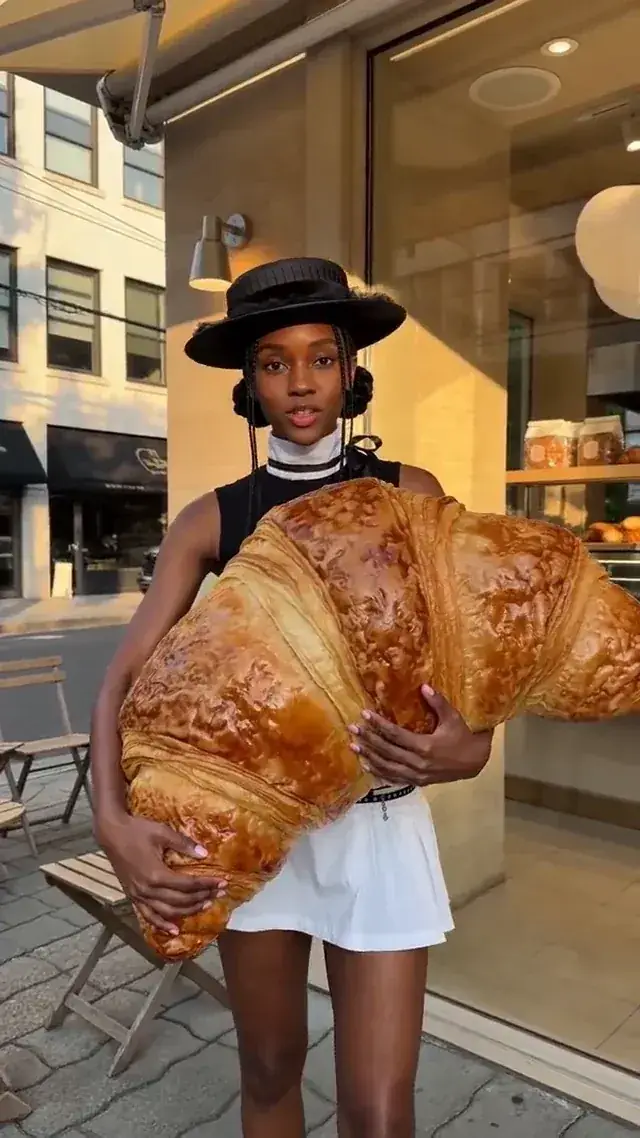 The same woman outside a bakery, hugging a giant croissant and talking to camera