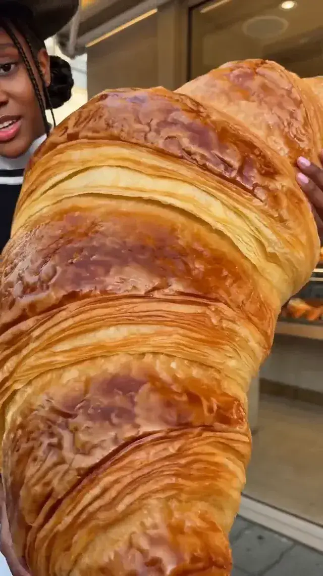 A creator holds an oversized croissant outside a bakery and speaks to camera